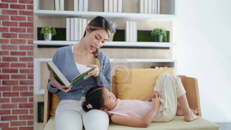 Photo for Happy Asian family mother reading book fairy tale story to daughter lying on sofa enjoying family lifestyle hobby at home. Happy family concept, Spending time together - Royalty Free Image