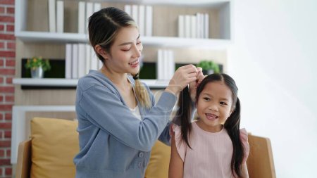 Téléchargez les photos : Heureuse famille aimante mère asiatique peigne les cheveux tressés de sa fille assis sur le canapé dans le salon. Traitement anti-poux - en image libre de droit