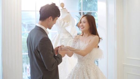 Téléchargez les photos : La belle mariée asiatique en robe de mariée et marié portant costume tenir la main au studio de mariage. Bon couple, jeunes mariés - en image libre de droit