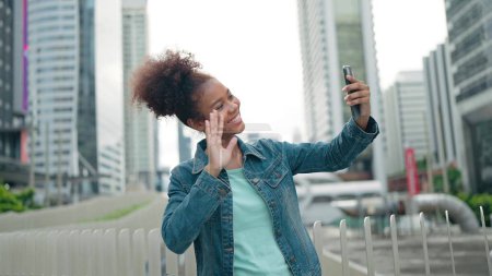 Foto de Hermosa joven turista de piel oscura hablando con amigos en el teléfono móvil mientras está de pie en la calle de la ciudad. Videollamada saludando mano con rascacielos modernos fondo - Imagen libre de derechos