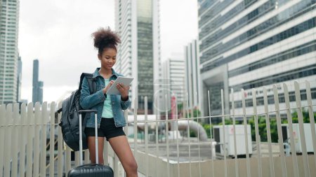 Téléchargez les photos : Jeunes femmes afro-américaines touristes debout dans le centre-ville en utilisant une tablette et souriant avec fond de gratte-ciel moderne - en image libre de droit