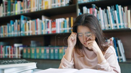 Téléchargez les photos : Asiatique femme étudiant portant des lunettes de lecture pour les devoirs et la préparation à l'examen dans la bibliothèque universitaire et sentir la douleur dans les yeux - en image libre de droit