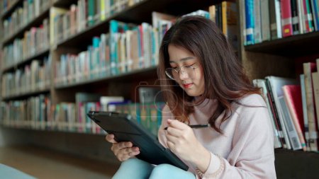 Téléchargez les photos : Femme asiatique portant des lunettes utilise une tablette assise dans la bibliothèque - en image libre de droit