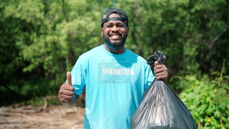 Téléchargez les photos : Noir afro-américain tenant sac poubelle et regardant la caméra, Volontaires nettoyage dans la forêt verte. Activités de RSE. Responsabilité sociale des entreprises. Problème environnemental. Journée de l'environnement - en image libre de droit