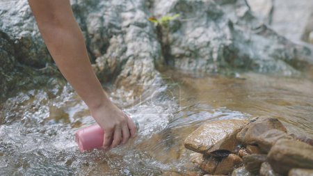 Téléchargez les photos : Main tenant une bouteille d'eau et puisant de l'eau dans la rivière ou la cascade de la forêt - en image libre de droit