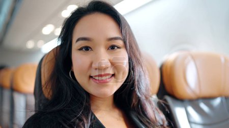 Foto de De cerca la cara de la hembra asiática mirando y sonriendo a la cámara en el avión, pasajero disfrutando de un vuelo cómodo mientras está sentado en la cabina del avión - Imagen libre de derechos
