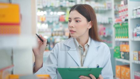 Téléchargez les photos : Professionnel asiatique femme pharmacien revérifie les produits en pharmacie et en utilisant le bloc-notes pour écrire des informations - en image libre de droit