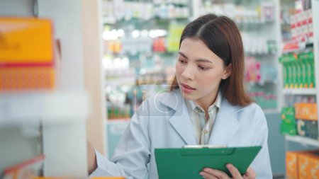 Photo for Professional Asian woman pharmacist rechecks products in pharmacy and using noteboard to write information - Royalty Free Image