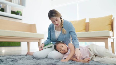 Téléchargez les photos : Mère asiatique lisant un livre et regardant petite fille dormir sur ses genoux dans le salon à la maison. Concept famille et amour - en image libre de droit