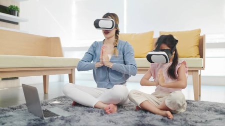 Photo for Asian mother and daughter wearing glasses of technology virtual reality headset doing yoga exercises in lotus position sitting on floor in living room. VR metaverse technology concept - Royalty Free Image