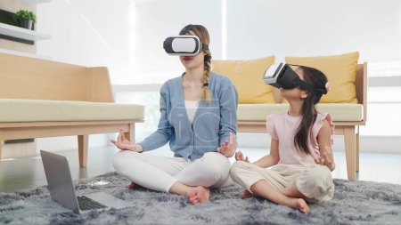 Téléchargez les photos : Mère et fille asiatiques portant des lunettes de technologie casque de réalité virtuelle faire des exercices de yoga en position lotus assis sur le sol dans le salon. Concept de technologie VR metaverse - en image libre de droit