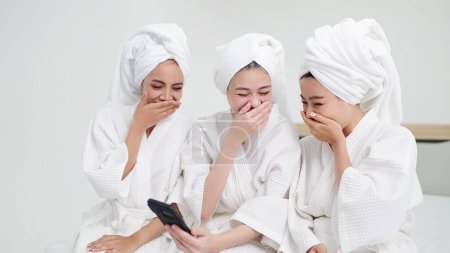 Téléchargez les photos : Trois jeunes belles femmes asiatiques en peignoirs et serviettes sur les têtes assises aiment bavarder et utiliser un smartphone. Meilleures amies en robes et serviettes - en image libre de droit