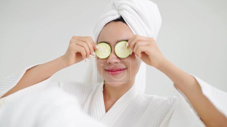 Photo for Close up face of beautiful young asian women in bathrobe and towels on head holding slices of fresh cucumber making refreshing eyes mask. Skin care concept - Royalty Free Image