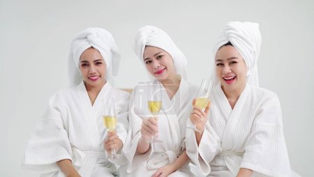 Foto de Tres jóvenes amigas felices en batas de baño y toallas en la cabeza sosteniendo copas de champán sonriendo a la cámara - Imagen libre de derechos