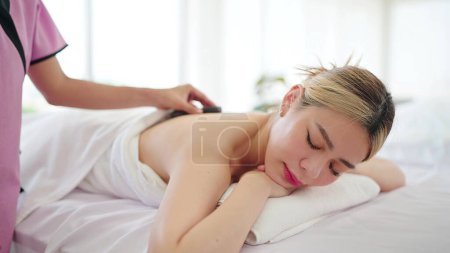 Photo for Close up face of beautiful Asian woman lying on massage bed while the masseuse is putting hot stones on back. Wellness and body relaxation therapy - Royalty Free Image