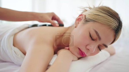 Photo for Beautiful Asian woman lying on spa bed with closed eyes and relaxed while masseuse putting hot stones on back. Spa back massage and wellness therapy - Royalty Free Image