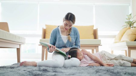 Foto de Asiática joven hermana leyendo un libro para niña mientras escucha en el regazo de la hermana en el suelo en la sala de estar. Concepto familia y amor - Imagen libre de derechos
