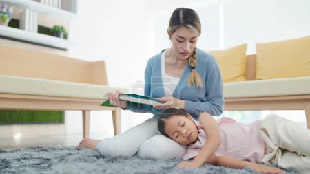 Foto de Madre asiática leyendo un libro y mirando a su hijita dormir en su regazo en la sala de estar en casa. Concepto familia y amor - Imagen libre de derechos