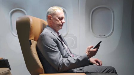 Foto de Hombre de negocios caucásico de mediana edad en traje sentado en el asiento en el avión utilizando el teléfono inteligente durante el vuelo de negocios y viajes - Imagen libre de derechos