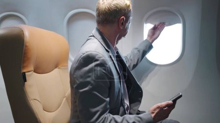 Téléchargez les photos : Homme caucasien d'âge moyen passager assis sur le siège dans l'avion fenêtre ouverte et regardant à l'extérieur tout en voyageant sur un long vol - en image libre de droit