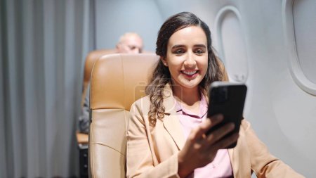 Foto de Feliz joven hispana latina pasajera sentada en el asiento usando un smartphone mientras viaja en avión. La mujer viaja en vuelo largo - Imagen libre de derechos
