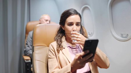 Photo for Hispanic latino businesswoman using smartphone and drinking while sitting in seat on airplane. Travel and business concept - Royalty Free Image