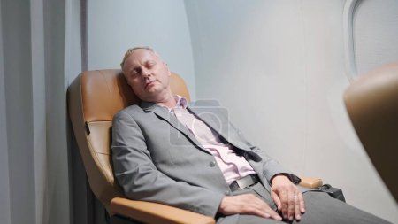 Foto de Exhausto hombre de negocios caucásico de mediana edad en traje durmiendo en el cómodo asiento de jet privado de negocios. El pasajero está durmiendo mientras viaja en avión - Imagen libre de derechos
