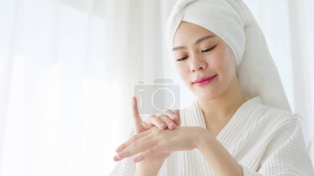 Photo for Asian woman in bathrobe and towel turban applying moisturizing cream after shower bath in the morning. Woman applying cream on hand. Skin care routine concept - Royalty Free Image