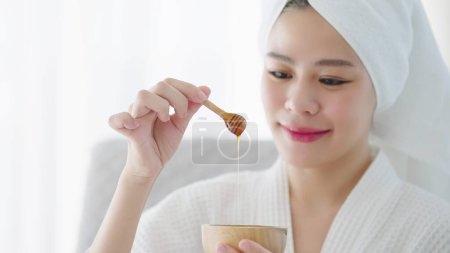 Téléchargez les photos : Gros plan main de la femme tenant cuillère à miel en bois goutte à goutte avec du miel pur dans un bol en bois et le fond est le visage bleui des jeunes femmes asiatiques. Soins de la peau spa relax concept - en image libre de droit