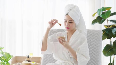 Foto de Hermosa mujer asiática joven en albornoz y turbante toalla sosteniendo un tazón de miel para el tratamiento de spa en casa el fin de semana. Concepto de belleza - Imagen libre de derechos