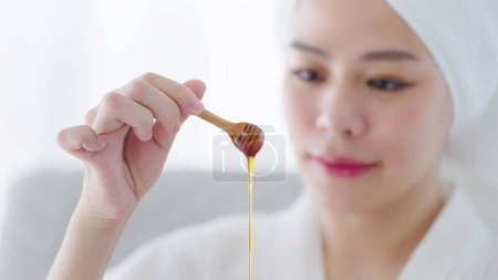 Photo for Blured background face of women. Close up hand of woman holding wooden honey scoop dripping with pure honey in wooden bowl for spa treatment. Skin care concept - Royalty Free Image