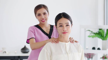 Téléchargez les photos : Asiatiques jeunes femmes et masseuse regardant caméra tout en se détendre pendant le massage des épaules assis dans le lit le week-end au spa station. Traiter la douleur avec massage - en image libre de droit