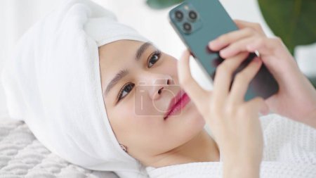 Téléchargez les photos : Gros plan visage de belle jeune femme asiatique portant une serviette sur la tête couchée sur le lit profiter de bavarder ou de vérifier les médias sociaux sur smartphone - en image libre de droit