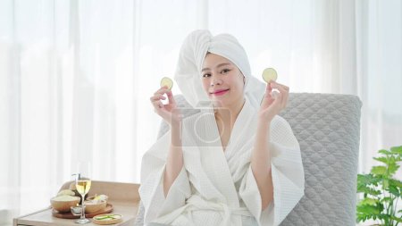 Téléchargez les photos : Belle jeune femme asiatique en peignoir et serviette turban tenant des tranches de concombre frais sur les mains souriant à la caméra tout en se relaxant sur le canapé-lit dans le salon moderne. Concept de spa beauté - en image libre de droit