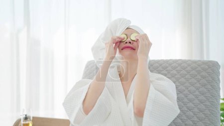 Foto de Mujer joven en albornoz y turbante toalla poniendo rodajas de pepino fresco en los ojos mientras se relaja en el sofá cama en la sala de estar moderna. concepto de spa de belleza - Imagen libre de derechos