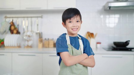 Photo for Confident asian little boy wearing apron smiling to camera whlie standing with arms crossed in kitchen room at home. Dream career concept - Royalty Free Image