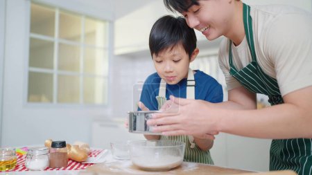 Foto de Asiático padre soltero enseñando a su hijo a tamizar la harina para cocinar o hornear pasar tiempo en vacaciones juntos en la sala de cocina en casa. Comida y cocina concepto, feliz familia asiática - Imagen libre de derechos