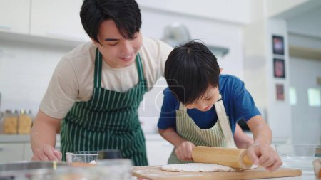 Téléchargez les photos : Père célibataire asiatique enseignant fils comment utiliser un rouleau à pâtisserie pour la cuisine ou la cuisson, passer du temps en vacances ensemble dans la salle de cuisine à la maison. Concept de famille heureuse - en image libre de droit