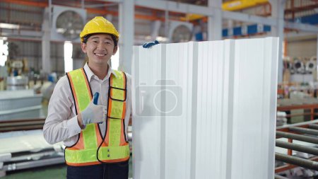 Téléchargez les photos : Portrait d'ingénieurs asiatiques en uniforme de sécurité tenant un toit en tôle couleur argent regardant la caméra tout en se tenant debout avec les pouces vers le haut à l'usine industrielle - en image libre de droit