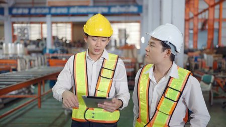 Téléchargez les photos : Deux ingénieurs asiatiques professionnels hommes en uniforme de sécurité tenant tablette de travail et de discuter tout en marchant à l'usine industrielle. Industrie manufacturière concept - en image libre de droit