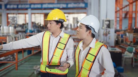 Téléchargez les photos : Deux ingénieurs asiatiques professionnels hommes en uniforme de sécurité tenant tablette de travail et de discuter tout en marchant à l'usine industrielle. Industrie manufacturière concept - en image libre de droit