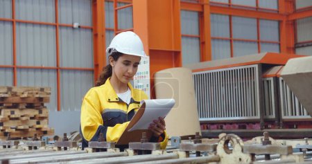 Photo for Young latin technician women in safety hard hat holding clipboard working and checking stock while walking at industrial factory - Royalty Free Image