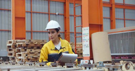 Téléchargez les photos : Jeune technicienne latine femmes en sécurité casque dur tenant presse-papiers de travail et de vérifier le stock tout en marchant à l'usine industrielle - en image libre de droit