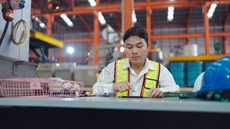 Téléchargez les photos : Jeune technicien asiatique hommes en uniforme de sécuritéde travailler sur tablette tout en se tenant à l'usine industrielle. Chatter ou envoyer un e-mail au gestionnaire par tablette - en image libre de droit