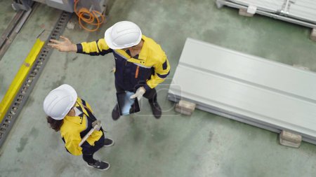 Foto de Vista superior de los ingenieros industriales hombres y mujeres que usan sombrero duro de seguridad de pie en la planta mientras trabajan y discuten juntos en la fábrica de fabricación de metal - Imagen libre de derechos