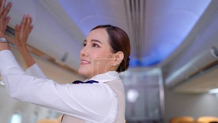 Téléchargez les photos : Des hôtesses de l'air asiatiques en uniforme fermant le compartiment à bagages supérieur dans l'avion. Enregistrement du coffre à bagages aérien avant le décollage de l'avion - en image libre de droit