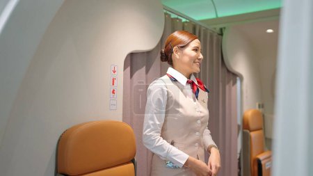 Téléchargez les photos : Belle hôtesse de l'air asiatique en uniforme travaillant dans l'avion. L'équipage de cabine debout à l'entrée de l'avion a souri amical et accueillant au vol. Transport aérien et tourisme concéder - en image libre de droit