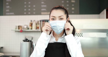 Photo for Portrait, Close up face of young woman barista in apron wearing surgical mask protective coronavirus covid19, global epidemic in coffee shop. Waitress wearing medical mask looking camera - Royalty Free Image