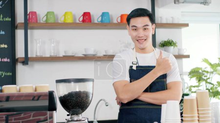 Foto de Retrato de atractivos hombres asiáticos jóvenes barista en delantal de pie con los brazos cruzados y pulgares hacia arriba sonriendo mientras mira a la cámara en el mostrador de la cafetería. Café concepto de propietario del negocio - Imagen libre de derechos