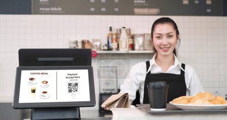 Foto de Mujer joven asiática camarera de pie en el mostrador trasero whlie poner bandeja con taza de café y pan, cruasanes en el mostrador en la cafetería. Menú y pago de código QR en monitor de pantalla en contador - Imagen libre de derechos